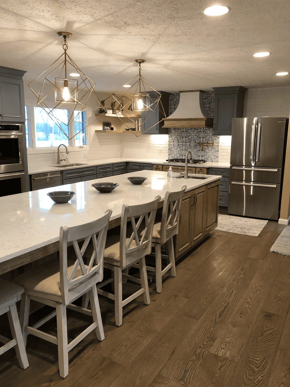 A modern kitchen with a large island, white countertops, wooden stools, stainless steel refrigerator, geometric pendant lights, and gray cabinets, featuring a patterned backsplash by the stove.