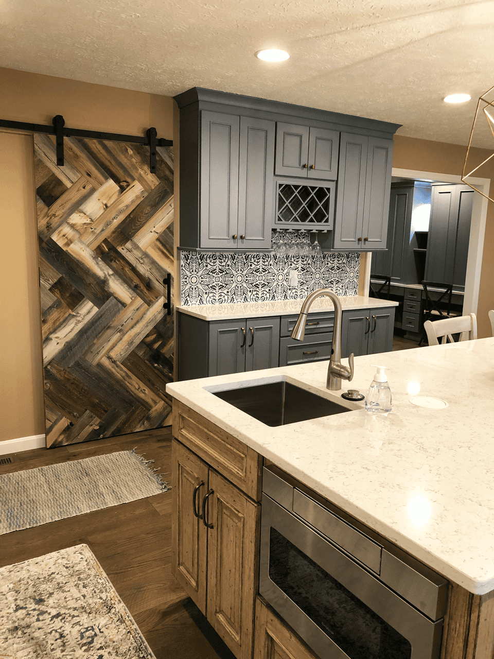 Modern kitchen with a central island featuring a sink and built-in oven. Grey cabinets above a patterned backsplash, and a sliding barn door to the side. Overhead lighting illuminates the space.