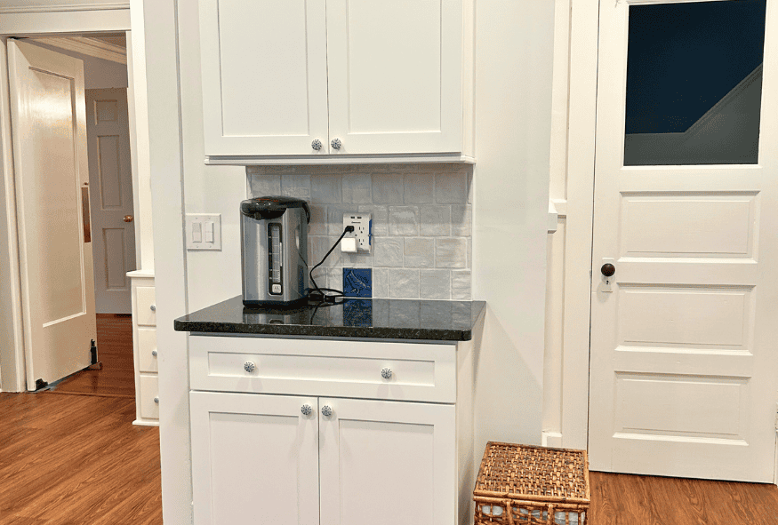 Oberlin kitchen remodel featuring white cabinets with a dark countertop, a coffee maker, tile backsplash, and a wicker basket on hardwood floors; white doors complete the clean, modern look.