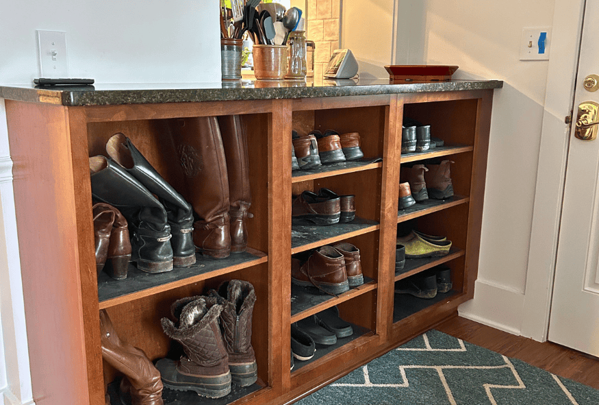 A wooden shoe rack with various boots, shoes, and slippers sits below a countertop holding kitchen utensils and other items, next to a door—perfect for your Oberlin kitchen remodel needs.