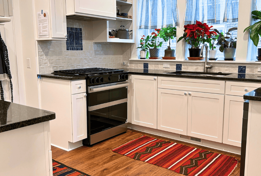 A modern Oberlin kitchen remodel featuring white cabinets, a stainless steel stove, a farmhouse sink, potted plants on the windowsill, and red patterned rugs on a wood floor.