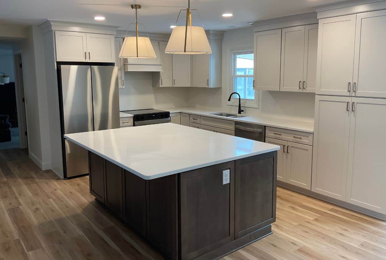 Modern kitchen remodel with white cabinets, stainless steel appliances, a large dark wood island with a white countertop, pendant lights, and light wood flooring—see the stunning Amherst Kitchen Remodel before and after transformation.