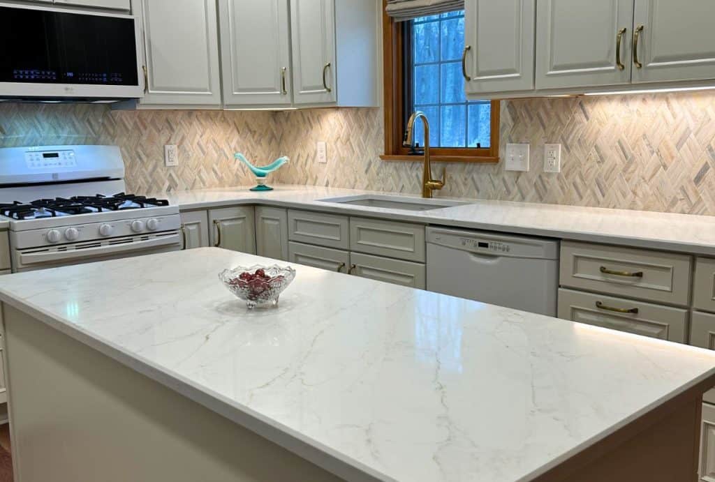 Modern Amherst Kitchen Remodel featuring white cabinets, marble countertops, gas stove, dishwasher, window above the sink, herringbone tile backsplash, and a glass bowl on the island—see the stunning kitchen before after transformation.