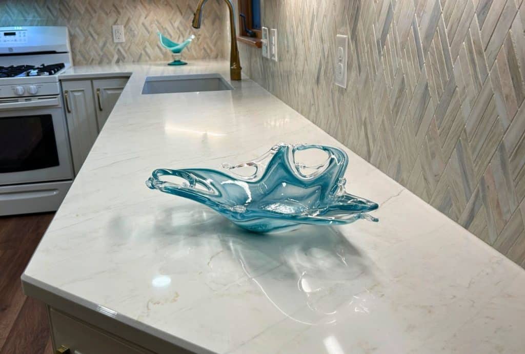 A blue glass decorative bowl sits on a white marble countertop near a sink, showcasing the elegant transformation of this kitchen remodel. In the background, a stove and another blue glass piece complete the updated look.