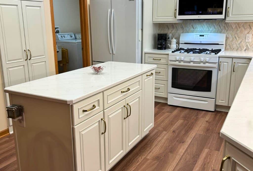 A modern Amherst Kitchen Remodel featuring white cabinets, marble countertops, a gas stove, refrigerator, microwave, island, and wood flooring. A laundry room is visible through an open doorway—see the stunning kitchen before after transformation.